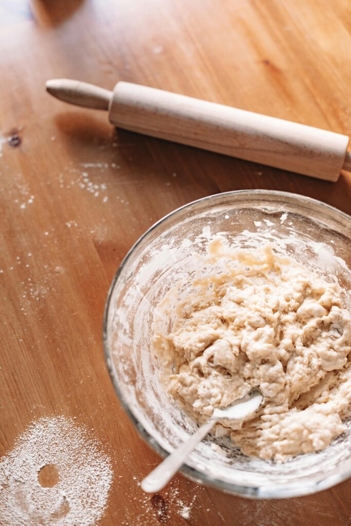 Close-up of a glass bowl with dough and a rolling pin on a wooden table.